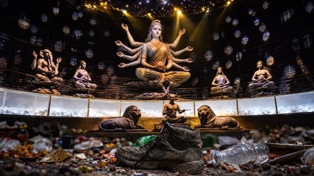 A mud-caked black shoe lies amid crushed plastic bottles, trampled marigolds, and debris on the floor of a circular bamboo-lattice Durga Puja pandal, while a golden-lit meditative idol of Goddess Durga and flanking deities sit untouched on a raised platform beneath spotlights in the background.