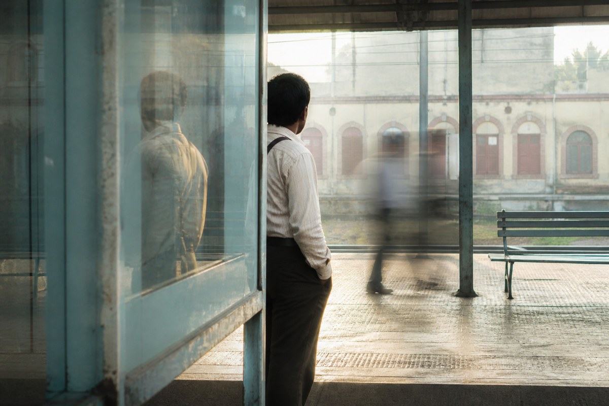 A commuter and a displaced reflection overlap in layered glass on a sunlit Kolkata railway platform