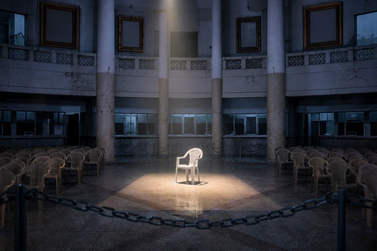 Single white chair stands spotlighted beyond chains in a neglected civic hall